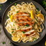 Creamy Lemon Chicken Pasta topped with juicy sliced grilled chicken, rich parmesan cream sauce, fresh parsley, and lemon wedges served on a dark plate.