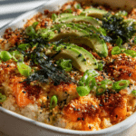 Easy sushi bake recipe in a baking dish topped with avocado slices, green onions, sesame seeds, and nori over seasoned sushi rice.