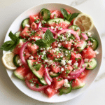 Fresh watermelon cucumber feta salad with red onion, mint, herbs, and lemon slices served in a white bowl.